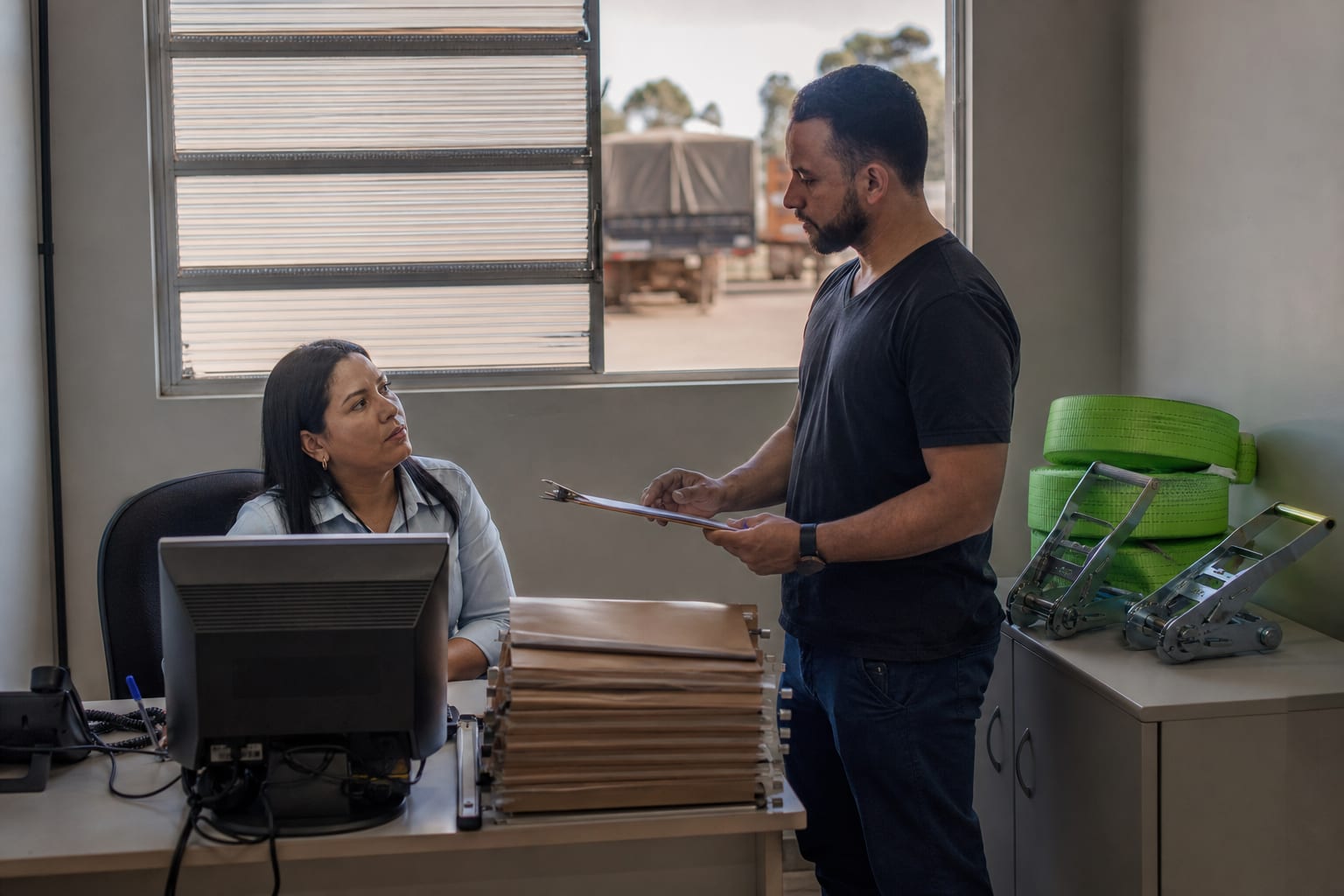 Dupla de funcionários conversa diante do computador em sala administrativa de transportadora do interior